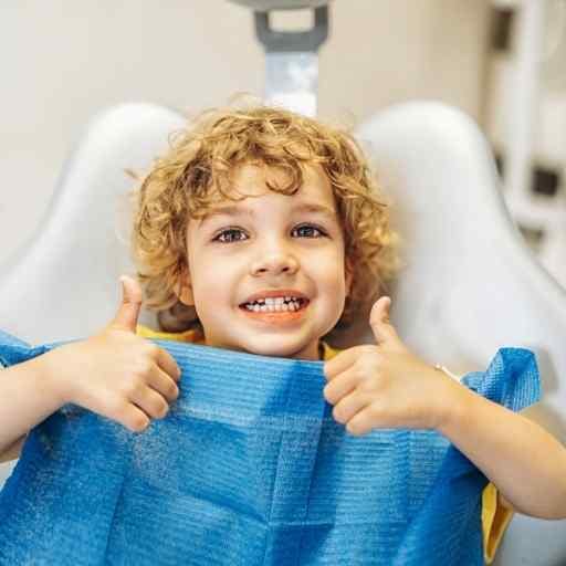 A young child with curly hair smiles brightly, giving a thumbs up while sitting in a dentist's chair, conveying joy and confidence in a dental setting.